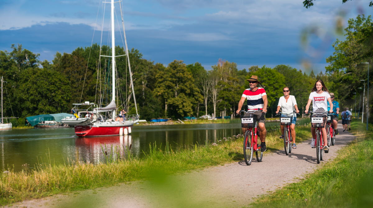 Sommarklädda personer cyklar längs Göta kanal och passerar en röd segelbåt i kanalen.