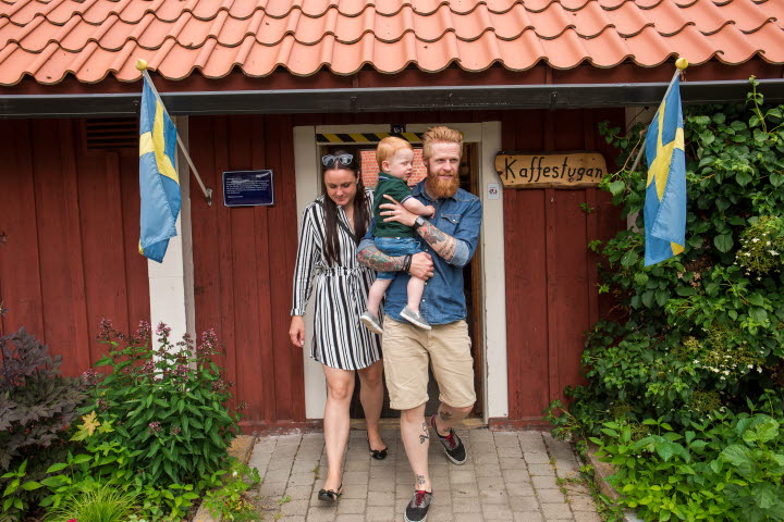 En man som håller ett barn i famnen och en kvinna går ut ur Kaffestugan på Turbinhusön i Tidaholm en sommardag.