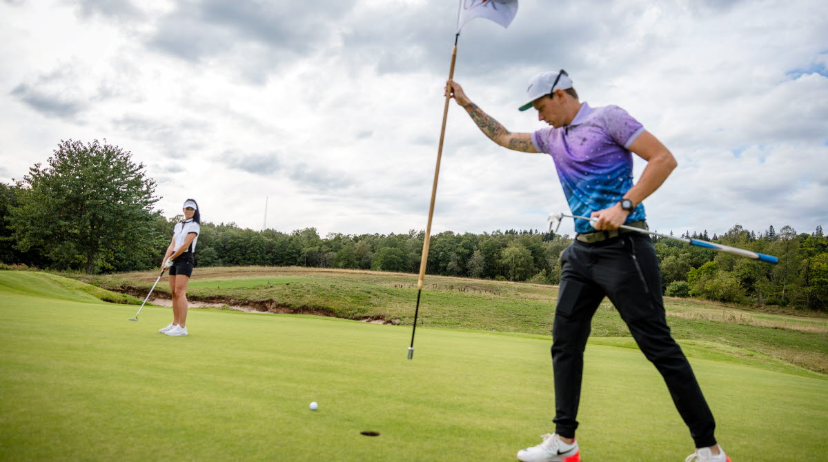 Två personer som golfar, killen i förgrunden håller i en golf flagga, tjejen i bakgrunden gör sig redo att slå
