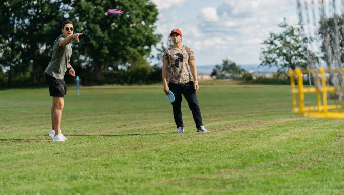 Två killar som kastar frisbee mot en gul korg i metall.
