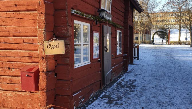 Fasaden på en röd timmerstuga. På knuten hänger en träskylt med texten "Öppet".