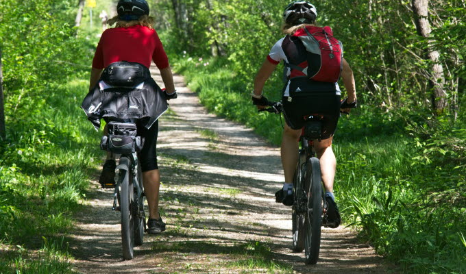 Två personer cyklar på en skogsväg. De bär ryggsäckar och hjälmar.