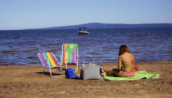 En flicka sitter på en handduk på en strand. Bredvid sig står två solstolar. Hennes vy är blått vatten och ett berg i horisonten.
