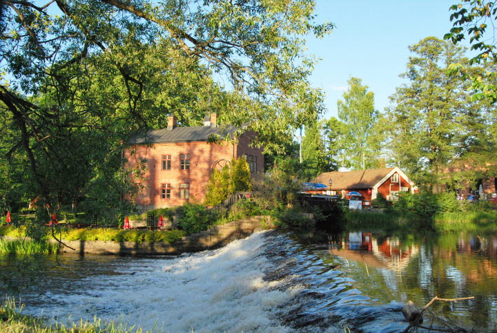 Bild på Turbinhuset som är Tidaholms konsthall och Kaffestugan med ett litet vattenfall framför i sommargrönska.