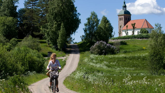 Kvinna kommer cyklandes på en grusväg med gröna åkrar runt om