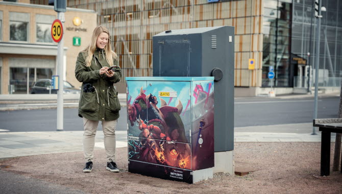 Kvinna i grön jacka och blont hår står med sin telefon i handen intill ett folierat elskåp som föreställer en spelkaraktär från spelet Battlerite.