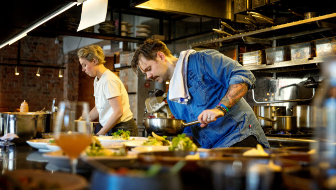 Två kockar står och lagar mat i en restaurang. Framför dem står tallrikar med mat på, färdiga att bli serverade.