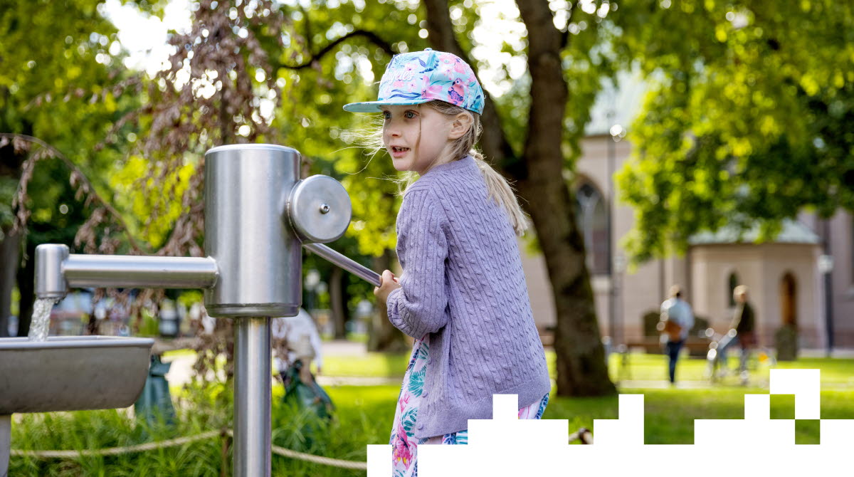 Flicka i färgglad keps och klänning med lila kofta över. Står och pumpar vatten ur en kran i en park. 