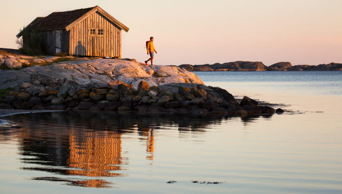 En person införd en gul tröja står på en klippa framför en gammal, grå sjöbod på Sydkoster. Havet ligger blankt och solen är på väg att gå ned.