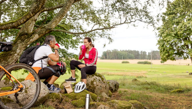 En kvinna och en man sitter vid kanten av ett fält och fikar.