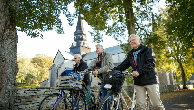 Tre män kommer gåendes och leder sina cyklar längs en stenmur. I bakgrunden ser man Varnhems klosterkyrka.