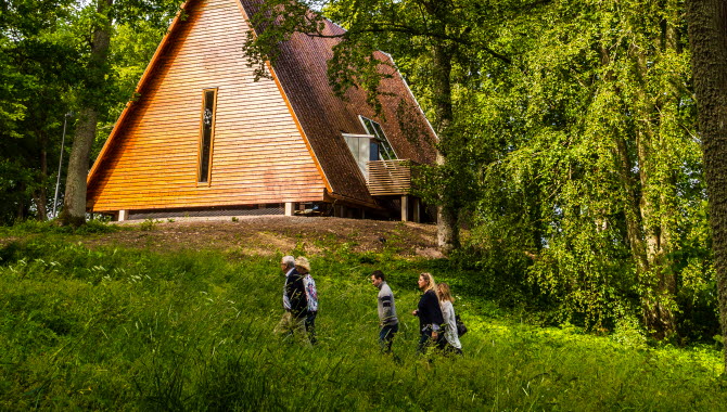 Kata gård, ett trähus på en kulle men grönskande träd omkring. Tre människor vandrar upp mot huset.