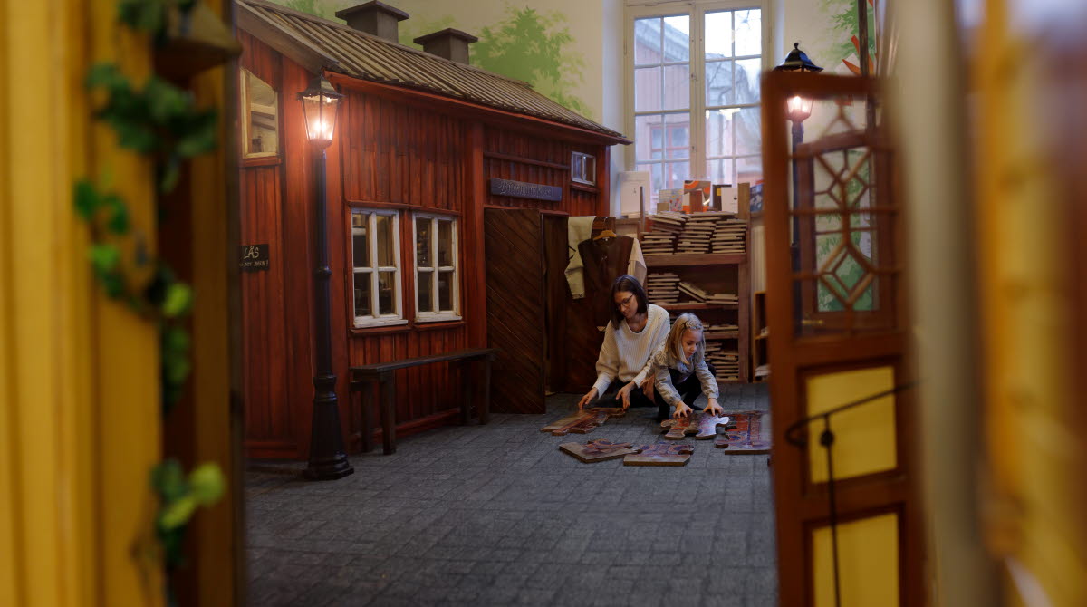 En vuxen och ett barn sitter på golvet framför ett äldre rödmålat hus i miniatyr inne i en utställning. 