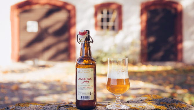 En produktbild på en öl och ett upphällt glas framör en välskött ladugård.