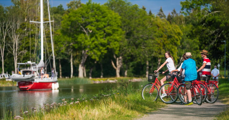 Tre personer stå med sina cyklar på kanalbanken och tittar på en segelbåt i Göta kanal.