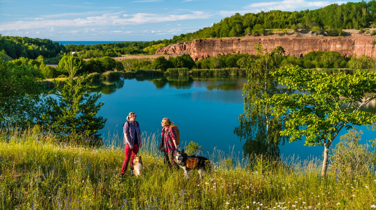 Två personer står vid kanten av ett vattenfyllt stenbrott med sina hundar.