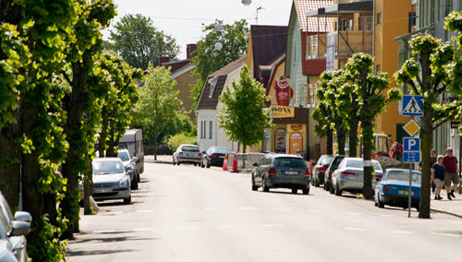 En somrig bild över en paradgata mitt i ett litet samhälle. Träden är gröna och bilar står parkerade längs gatan.