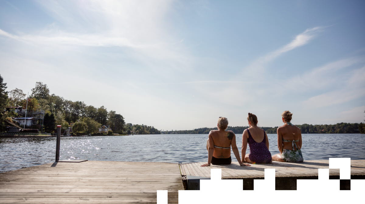 Tre kvinnor i badkläder sitter med ryggen mot kameran på en brygga och blickar ut över en sjö. 